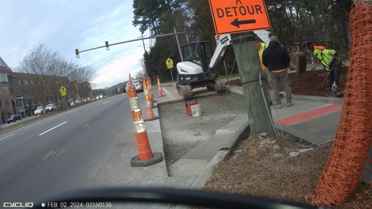 Screengrab from a bike cam video showing a construction crew installing a bulb out and new crosswalk where a side street intersects with an arterial. There is a detour sign directing pedestrians from the sidewalk across the arterial.