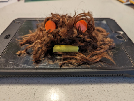Close-up photo of the face of a monster, made from a pile of recently-shorn dog hair, with orange ear plugs for eyes and a piece of yellow chalk as a mouth. The monster, named Peterson, is sitting on top of a small black ipad.