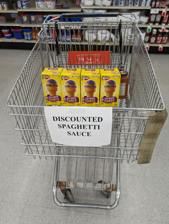 Photo of a shopping cart with a sign on the front reading, "Discounted Spaghetti Sauce." In the cart is a case of ice cream cones.
