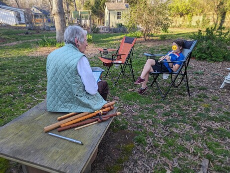 Photo showing an older woman sitting outside on a wooden table, surrounded by wooden flutes, recorders, and penny whistles. A child lounges nearby in a lawn chair, playing a white plastic recorder