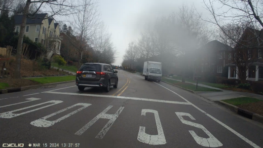 Screengrab from a bicycle handlebar camera (so, POV is the cyclist) showing a street, with the word "SCHOOL" painted in large white letters across the street, indicating a school zone in the foreground. In the near distance, a white landscaping truck is parked in the bike lane, facing the wrong direction. There is also an SUV with a baby-on-board sticker passing the cyclist.
