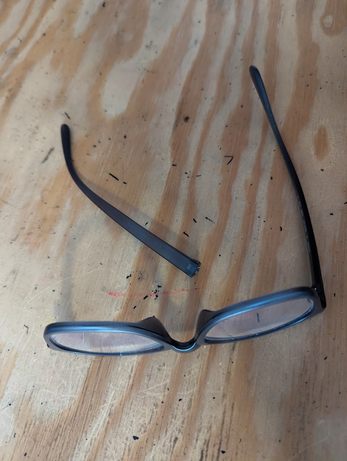 Photo of a pair of glasses, with one arm broken off, sitting on the surface of a well-used wooden desk.